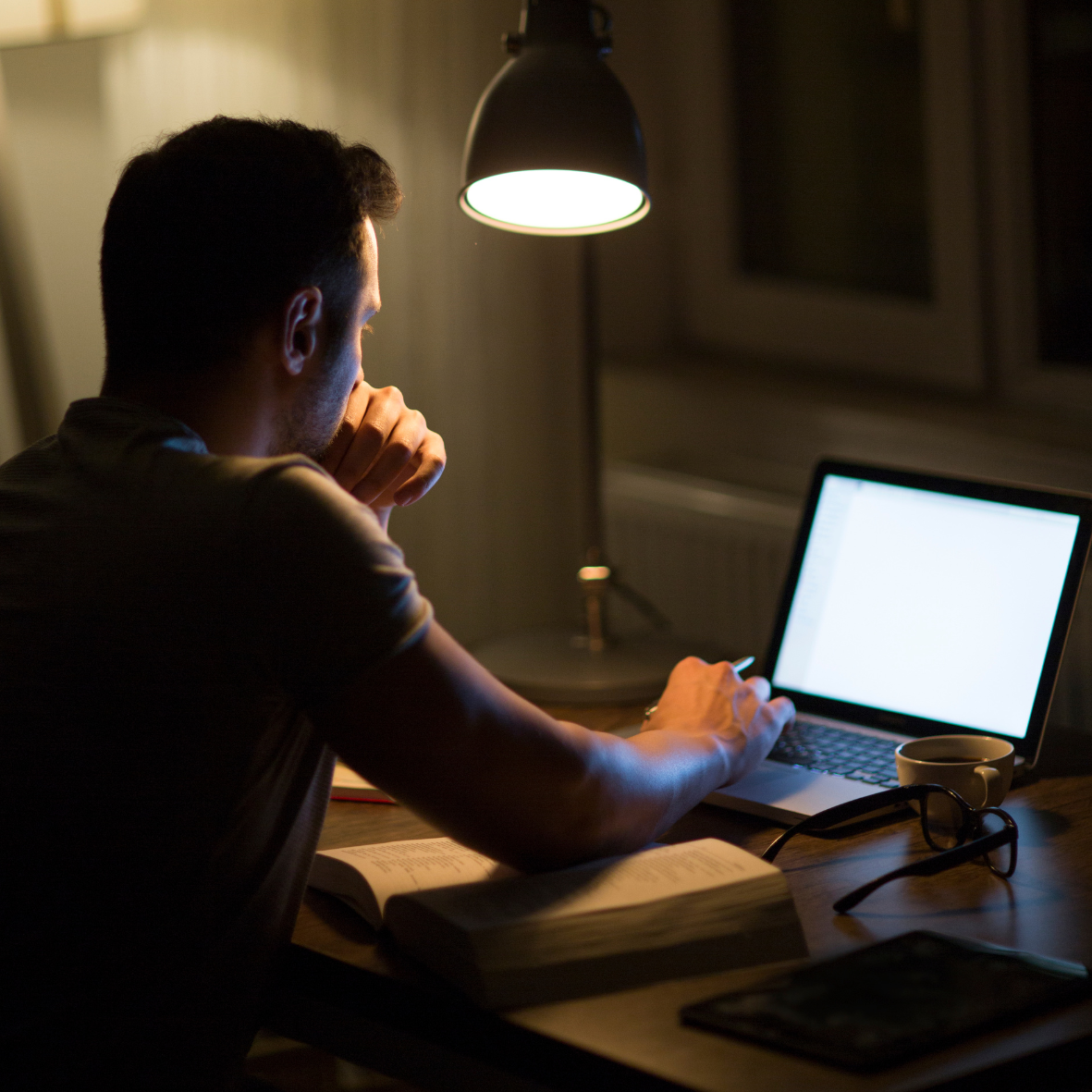 Focused man working late at night at his desk with a laptop and open book, lit by a single desk lamp—symbolising dedication and the grind behind building a business.