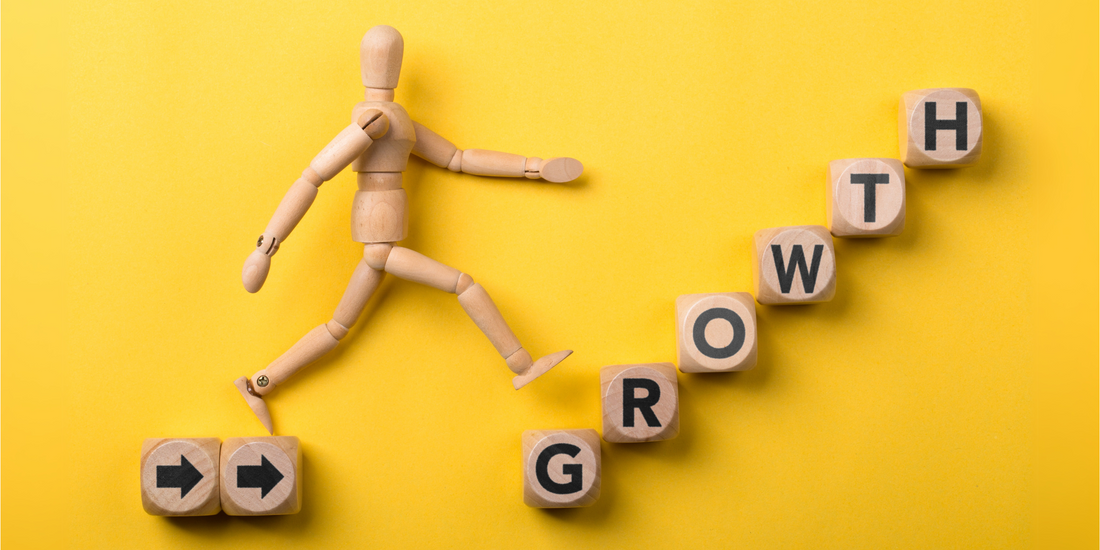 A wooden mannequin figure stepping upward on wooden blocks spelling out the word "GROWTH" against a vibrant yellow background, symbolising progress and business development.