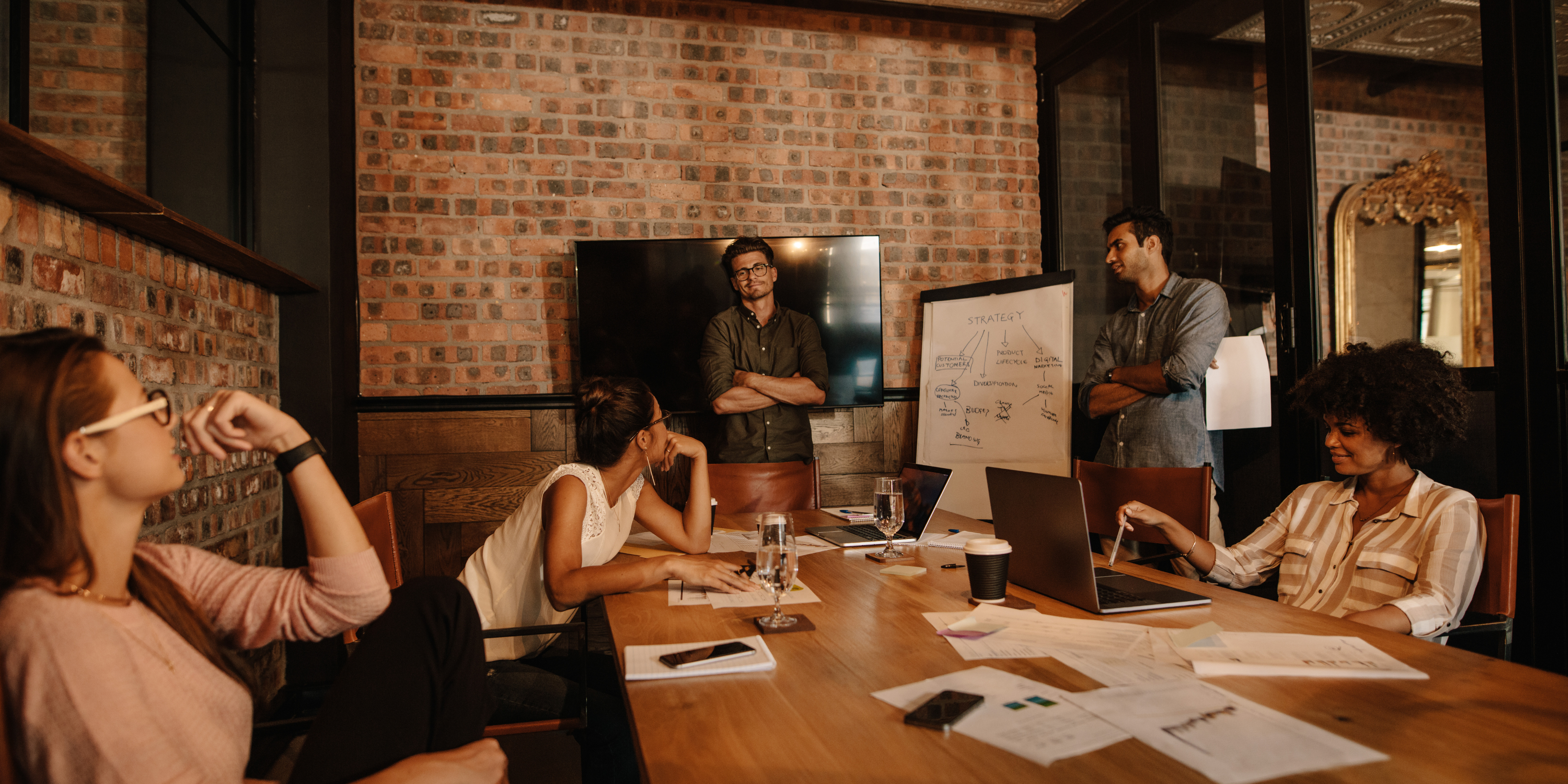 Diverse group of young professionals in a strategy meeting, gathered around a table with charts, laptops, and whiteboard notes, symbolising startup planning and collaboration.
